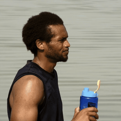 En mand i ærmeløs skjorte står udendørs og holder en blå vandflaske med et gult sugerør. Han kniber øjnene sammen i sollyset, og hans hud ser svedig ud, hvilket tyder på, at han har trænet.