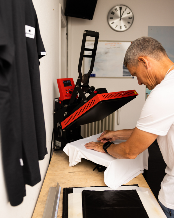 En person betjener en varmepressemaskine for at printe et design på en hvid t-shirt på et arbejdsbord. I baggrunden ses et ur og en hængende sort t-shirt.