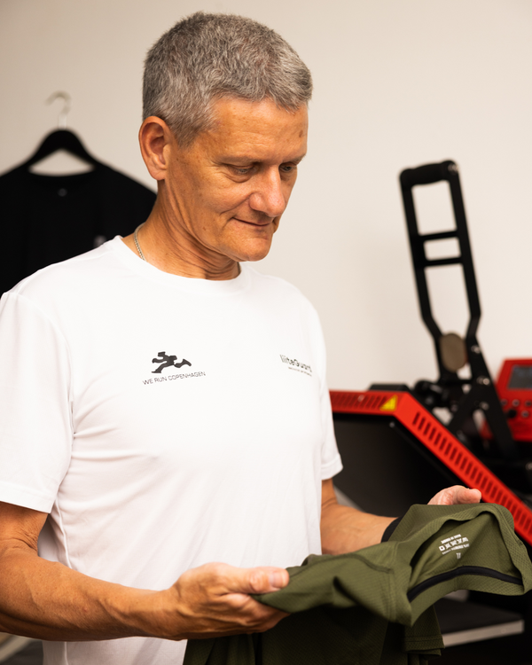 En mand med kort gråt hår iført hvid T-shirt står indendørs med en foldet olivengrøn skjorte. I baggrunden er der en rød og sort maskine og en mørk skjorte, der hænger på en krog.