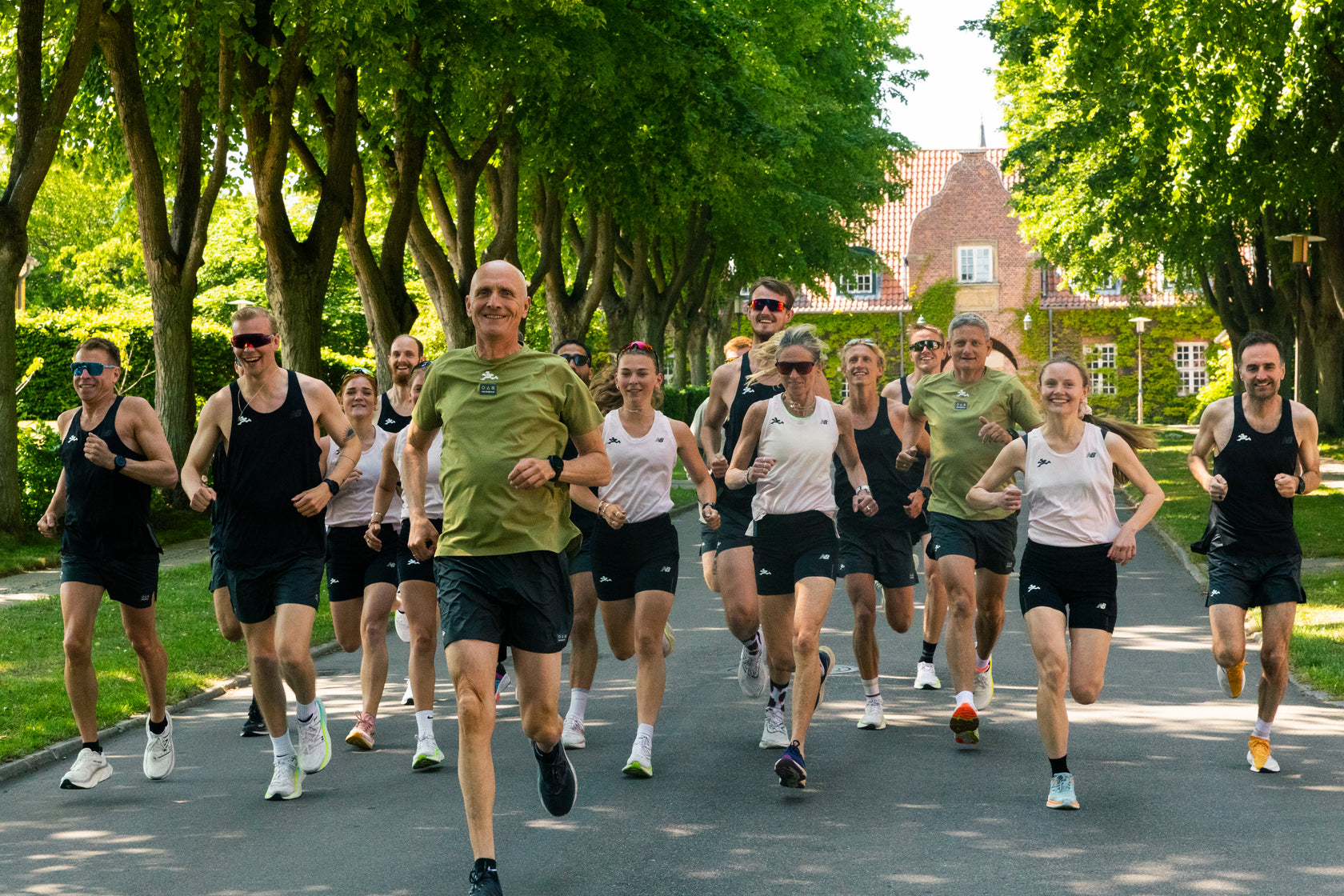 En gruppe mennesker jogger sammen udendørs på en gade med træer i klart dagslys. De fleste er iført sportstøj og ser ud til at smile eller hygge sig. Et murstenshus er synligt i baggrunden.