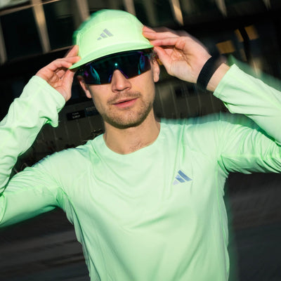 En mand iført neongrøn langærmet skjorte, matchende kasket og mørke solbriller justerer sin hat, mens han står udendørs. Adidas-logoet er synligt på hans skjorte og kasket.