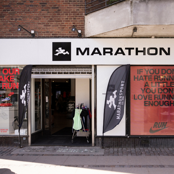Butiksfacade til en butik, der hedder Marathon, med to sorte flag, der viser butikkens logo. Indgangen er åben og afslører sportstøj indenfor. Et motiverende citat er trykt på det højre vindue.