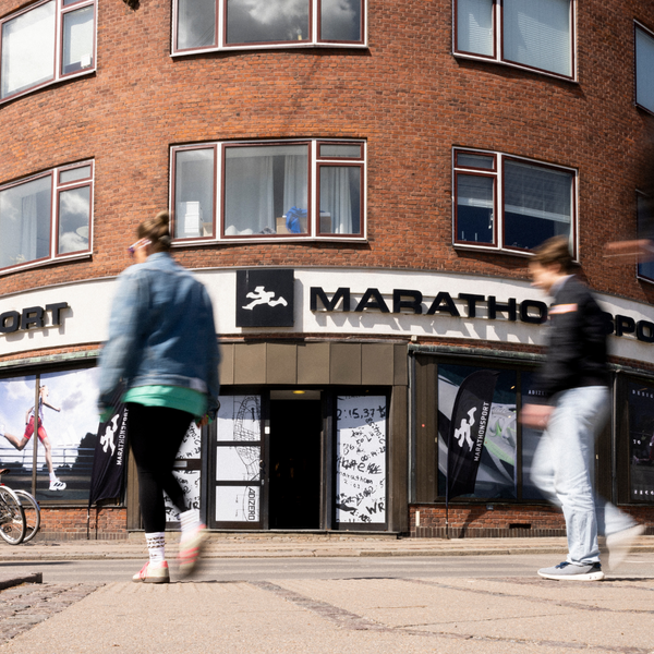 To personer går forbi indgangen til en butik ved navn MARATHON SPORT i stueetagen af en buet murstensbygning med flere store vinduer. En cykel er parkeret til venstre for butikken.