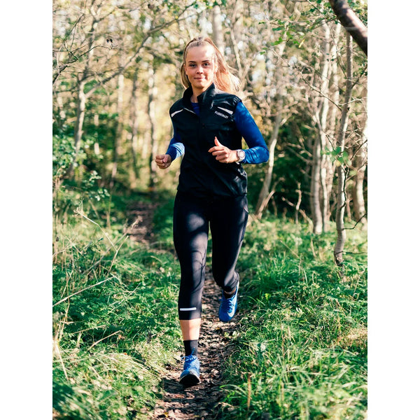 En person iført Fusion 3/4 Training Tights, Dame, SuperBlack og blå sko jogger ad en smal grussti gennem et skovområde med grønt græs og træer.