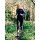 En person iført Fusion 3/4 Training Tights, Dame, SuperBlack og blå sko jogger ad en smal grussti gennem et skovområde med grønt græs og træer.
