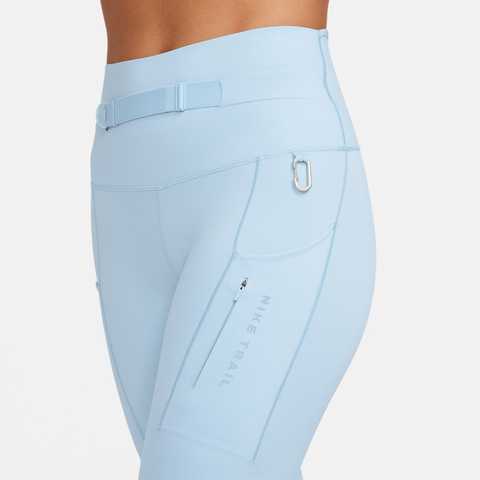 Nærbillede af en person iført Nike Trail High-Waisted 7/8 Leggings til kvinder i Lt Armory Blue/Lt Orewoord Brn/Khaki, med bælte, lynlåslomme, karabinhage og "Nike Trail"-tekst på låret mod en hvid baggrund.