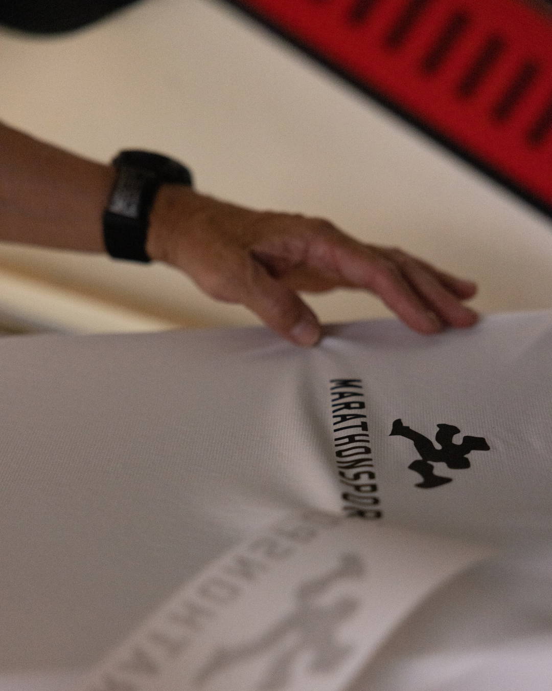 En person iført et sort armbåndsur rører ved en hvid beklædningsgenstand med et sort logo og ordet KARATEKNISSPAC. Et rødt og sort objekt er delvist synligt i baggrunden.