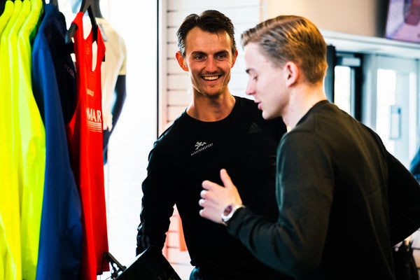 To mænd står indendørs i nærheden af en udstilling med farvestrålende sportstrøjer. Den ene mand smiler, mens han lytter til den anden, som taler og bærer et ur. Baggrunden består af hylder og et vindue.