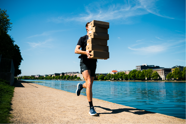 En person i sportstøj jogger på en sti ved siden af et vandløb, bærende på en høj stak pap-pizzabakker, der delvist skjuler ansigtet. Bygninger og træer står på den modsatte side af vandet under en klar blå himmel.