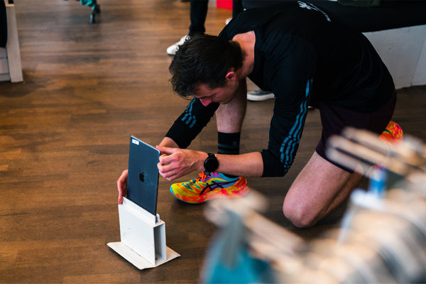 En person knæler på gulvet og justerer en iPad, der er placeret lodret i et hvidt stativ, iført en sort langærmet skjorte, shorts og farverige løbesko, indendørs på et trægulv.
