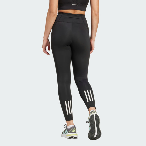 En person iført sorte Adidas OTR 7/8 Tights, Dame, parret med en sort sports-bh og gråhvide sportssko med grønne detaljer, går sin vej. Strømpebukserne har tre hvide, lodrette striber nær læggene.