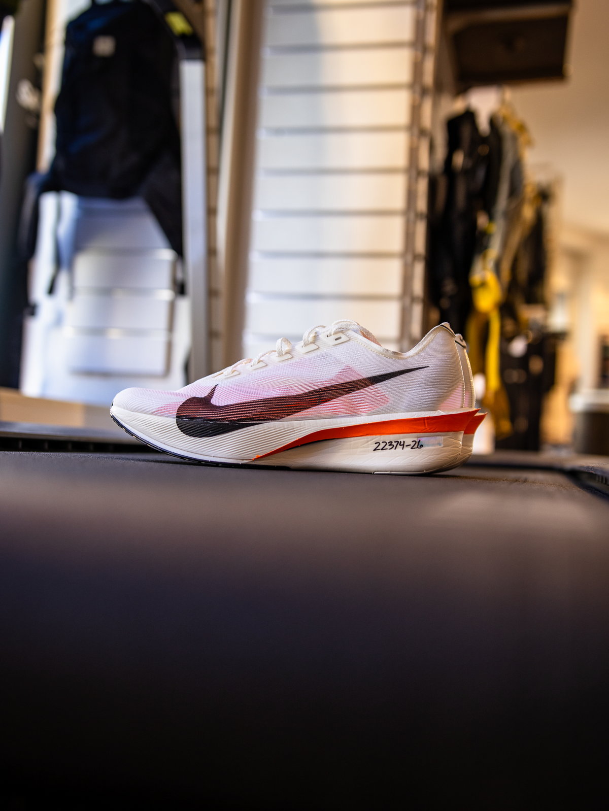 En hvid Nike-løbesko med sort swoosh og røde detaljer er udstillet på en sort overflade i en butik, hvor hylder og andre varer er synlige i den slørede baggrund.