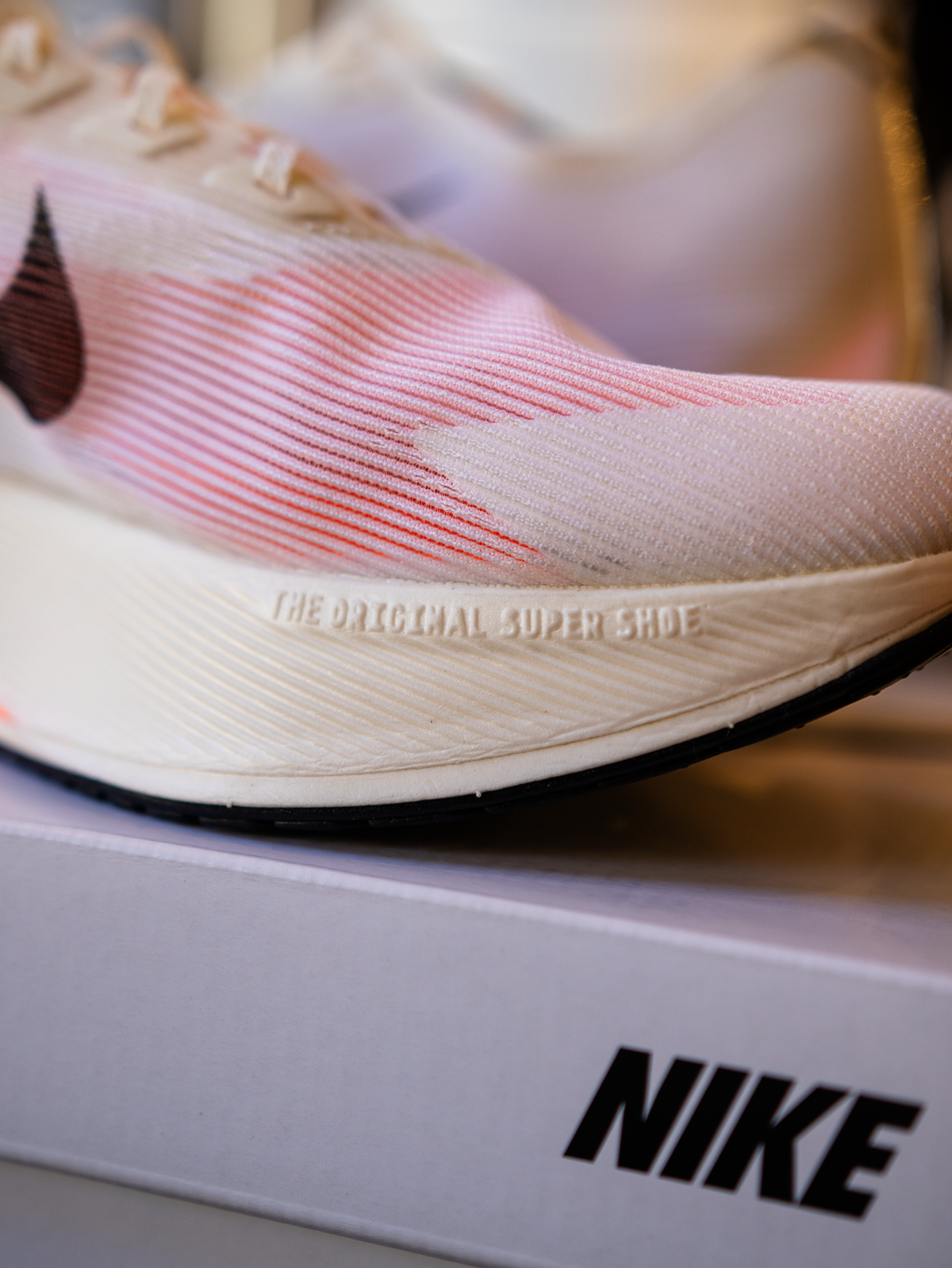 Nærbillede af en hvid Nike-løbesko med pink og røde detaljer, placeret oven på en hvid Nike-æske. Skoen har teksten THE ORIGINAL SUPER SHOE præget på siden af sålen.