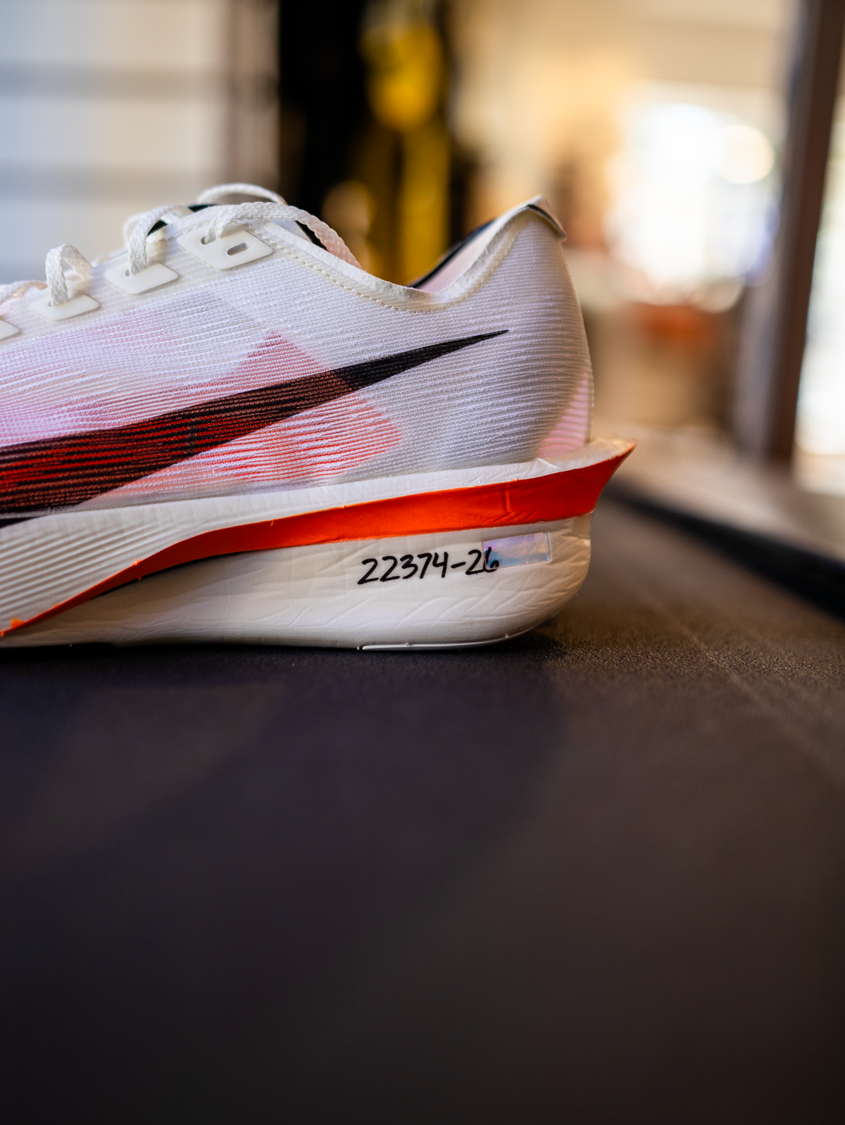 Et nærbillede af en hvid løbesko med et rødt og sort swoosh-design. På hælen ses de håndskrevne tal 22374-26 med sort blæk på en rød baggrund. Skoen hviler på en mørk overflade indendørs.