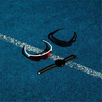 To par sportssolbriller og et sort smartwatch er placeret på en blå løbebane med en hvid linje, der løber diagonalt gennem midten af billedet.