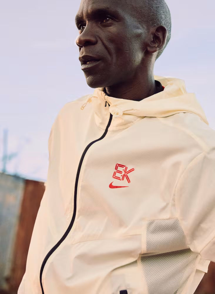 En mand iført en lys jakke med lynlås og rødt EK- og Nike-logo står udendørs. Jakken har en hætte og en sort lynlås. Baggrunden er ude af fokus med en synlig struktur.