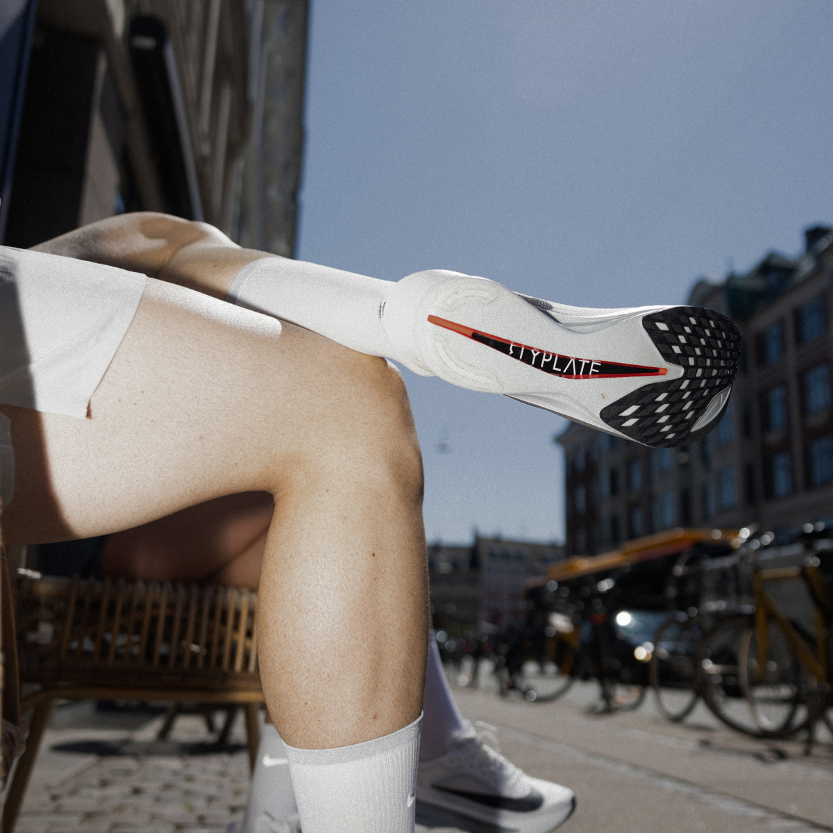 En person sidder udendørs med det ene ben krydset over det andet, iført hvide sokker og hvide sportssko, hvor der står FLYPLATE på sålen. Cykler og bygninger er synlige i baggrunden.