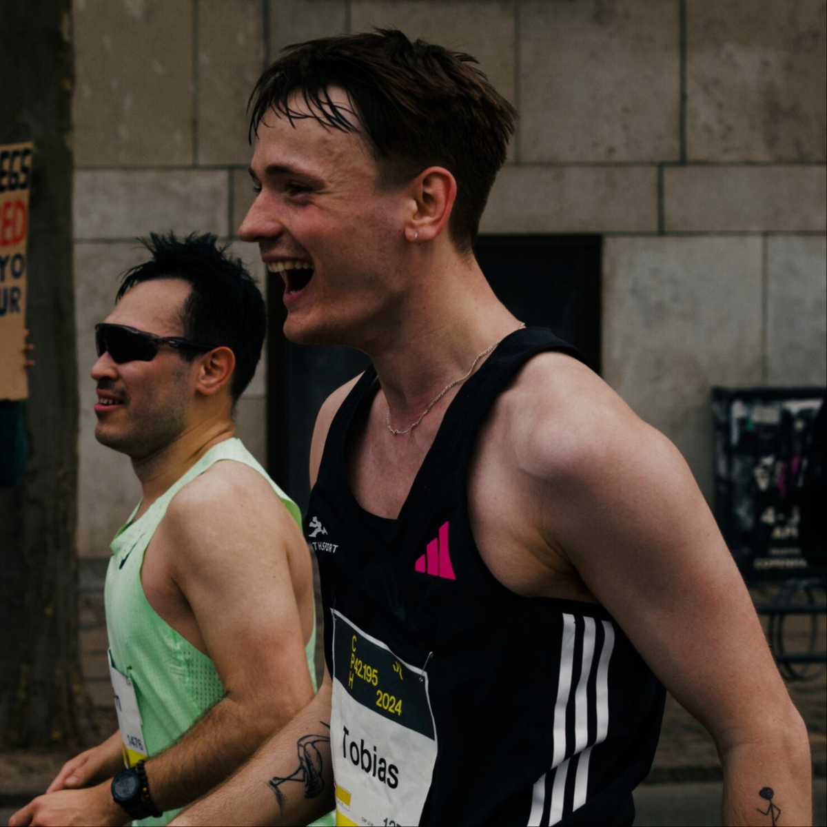 To løbere deltager i et løb, den ene i en sort tanktop og den anden i en lysegrøn tanktop, begge har startnummer på og løber udendørs i nærheden af en betonmur.