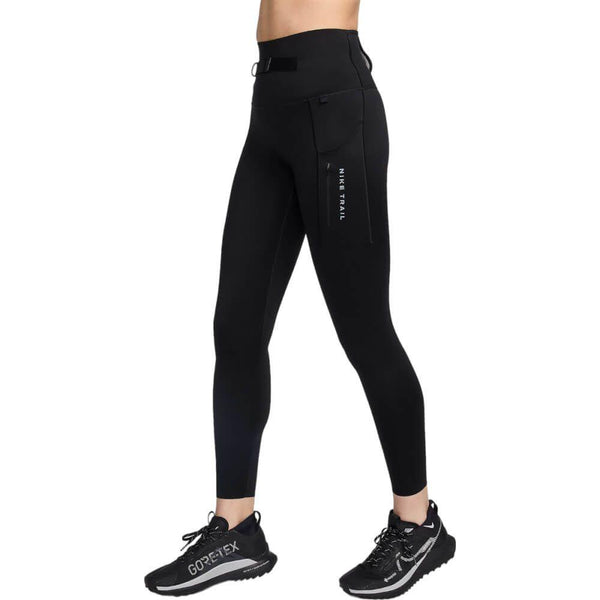 En person er iført sorte Nike Trail High-Waisted 7/8 Leggings (Dame, Black/Dk Smoke Grey) med Dri-FIT-stof og NIKE TRAIL-tekst på lommen, sammen med sorte GORE-TEX-trailløbesko, på en almindelig hvid baggrund.