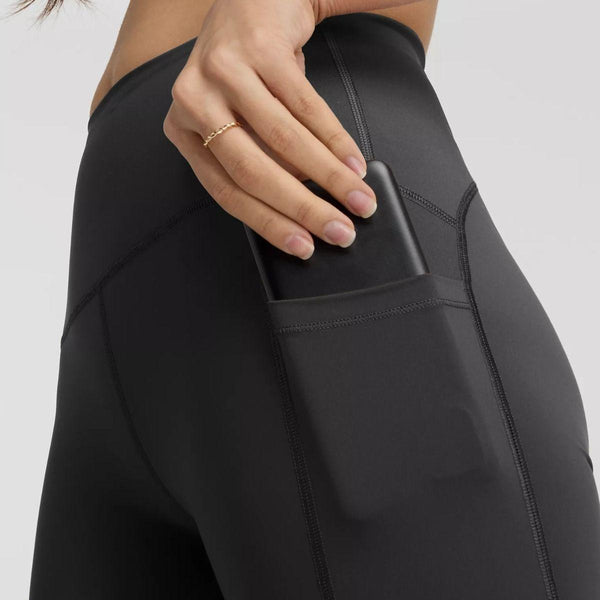 En person iført New Balance Sleek Pocket High Rise Tights, Dame, Black stikker en smartphone ned i tightsens sidelomme. Kun underkroppen og en hånd med en tynd guldring er synlige.