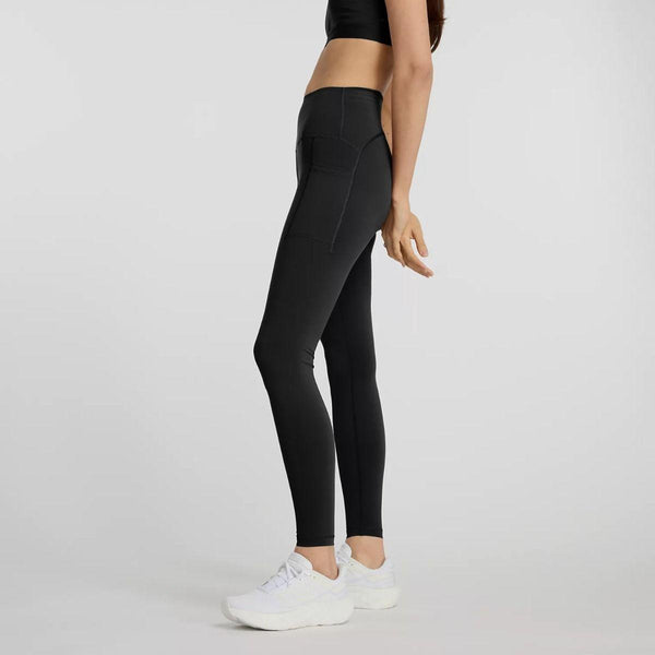 En kvinde i sideprofil bærer New Balance Sleek Pocket High Rise Tights, Dame, Black - sorte højtaljede leggings med sidelommer og en matchende sports-bh - sammen med hvide sportssneakers på en lys baggrund.