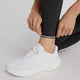 En person justerer manchetten på sorte New Balance Sleek Pocket High Rise Tights til kvinder, parret med hvide sneakers. Kun underbenene og hænderne er vist mod en ensfarvet baggrund.