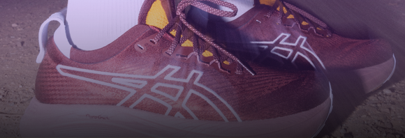 Nærbillede af en person iført rødbrune ASICS-løbesko med hvide såler og logo, der står på en jordoverflade. Personen har også hvide sokker på.