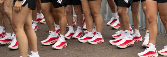 En gruppe mennesker iført matchende sorte shorts, hvide sokker og hvide sneakers med røde detaljer står sammen på en asfalteret plads i nærheden af et metalhegn.