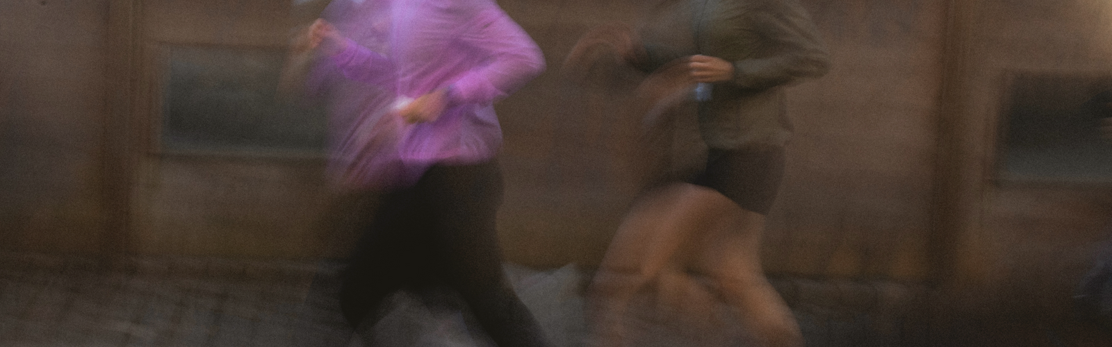 To personer i bevægelsessløring jogger side om side udendørs, den ene iført lilla top og sorte bukser, den anden i mørk jakke og shorts. Baggrunden er ude af fokus.