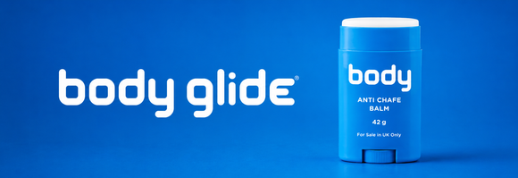 En blå Body Glide anti-chafe balm stick ligger på en blå overflade ved siden af en stor hvid tekst, hvor der står body glide på en ensfarvet blå baggrund.