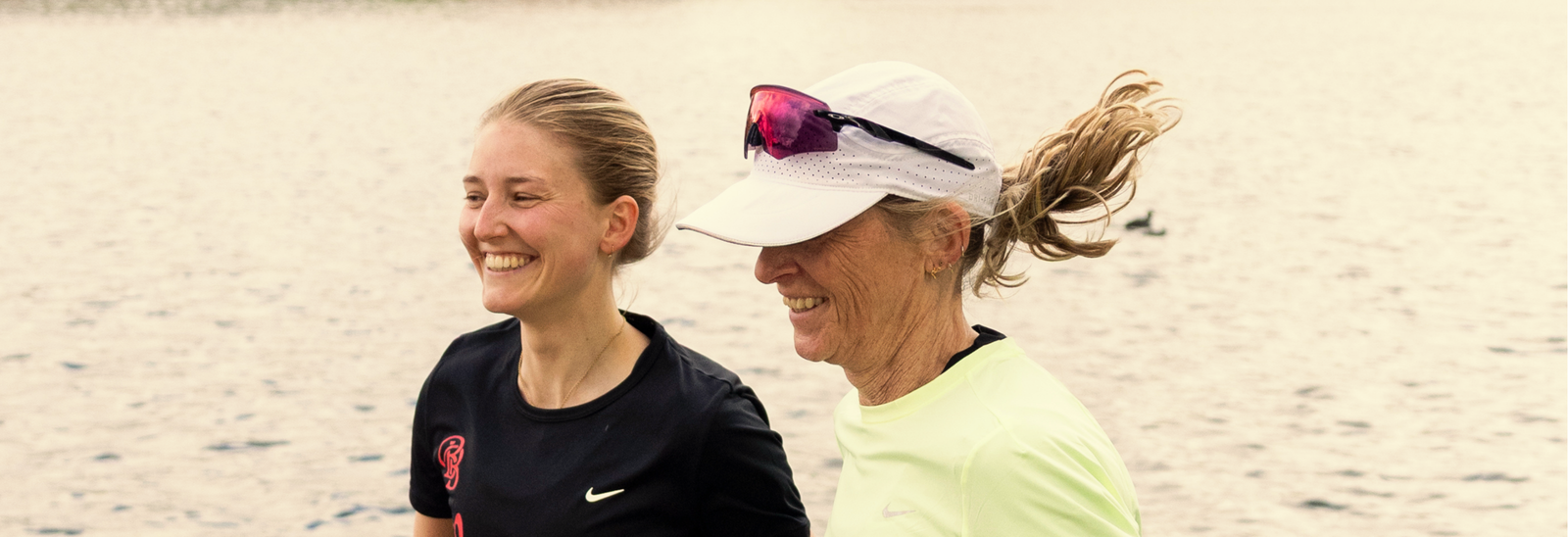 To kvinder jogger side om side i nærheden af et vandløb. Den ene har en sort skjorte på og smiler, mens den anden har en hvid kasket, solbriller og en lysegul skjorte på. Begge ser tilfredse og afslappede ud.