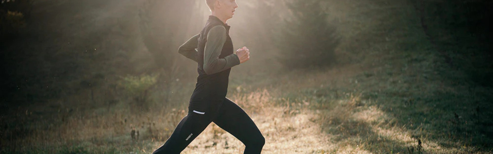 En person iført sportstøj løber udendørs på en græsklædt sti med sollys, der strømmer gennem træer i baggrunden.