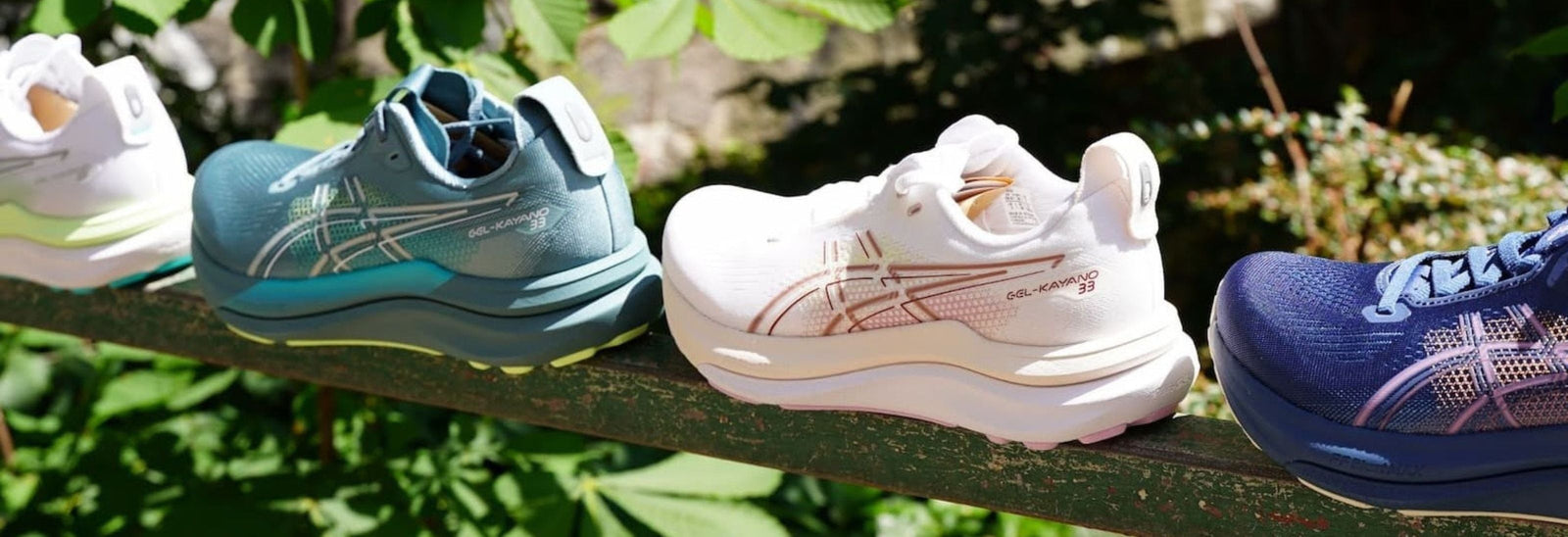 Fire par ASICS-løbesko i blå, hvide og marineblå farver er stillet op side om side på et metalgelænder udendørs med grønne planter i baggrunden.