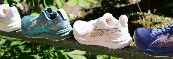Fire par ASICS-løbesko i blå, hvide og marineblå farver er stillet op side om side på et metalgelænder udendørs med grønne planter i baggrunden.