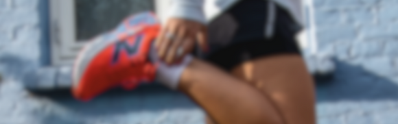 En person i sportstøj strækker sit ben og holder om anklen med den ene hånd. Den orange sneaker med et blåt N-logo er fremtrædende. Baggrunden viser en blå væg og et vindue. Billedet er sløret.