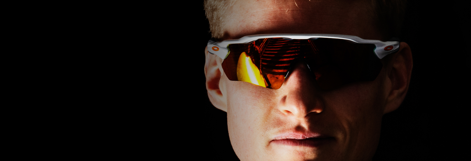 Nærbillede af en person iført orangefarvede sportssolbriller med hvidt stel på en mørk baggrund. Kun en del af personens ansigt er oplyst og synligt.