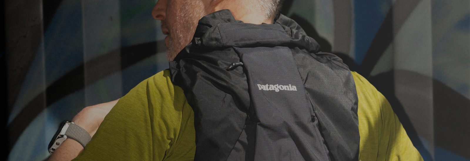 En person iført en grøn skjorte og en sort Patagonia-rygsæk ses bagfra. Personen har også et smartwatch på sit venstre håndled. Baggrunden er et abstrakt vægmaleri.