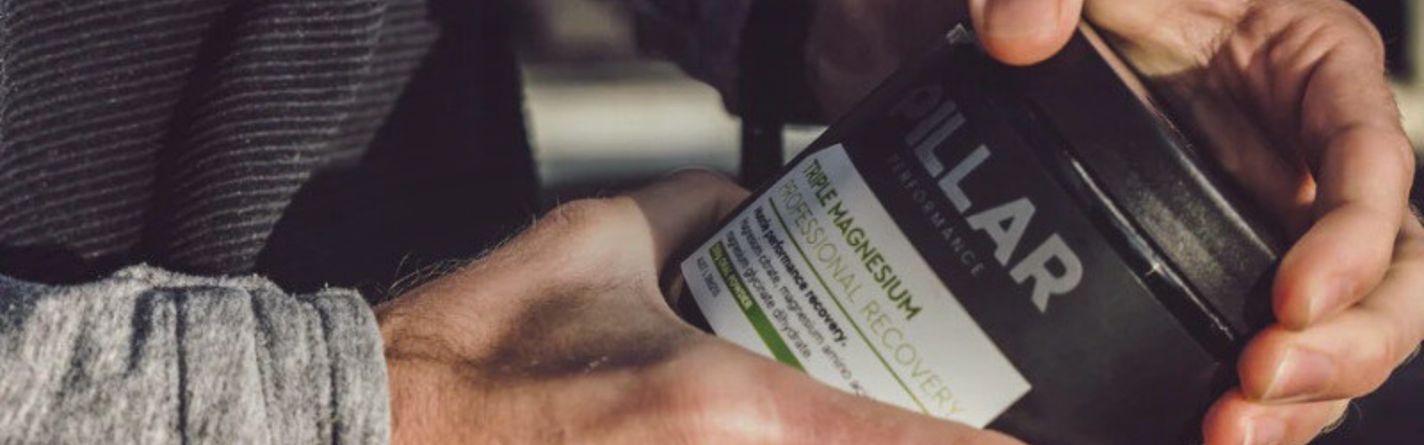 En person i en grå, langærmet skjorte åbner en sort beholder mærket "Pillar" med en tekst, der nævner magnesium og recovery. Beholderen holdes med begge hænder.
