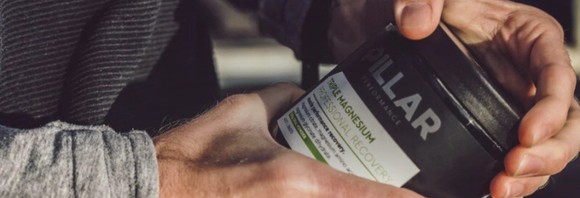 En person i en grå, langærmet skjorte åbner en sort beholder mærket "Pillar" med en tekst, der nævner magnesium og recovery. Beholderen holdes med begge hænder.