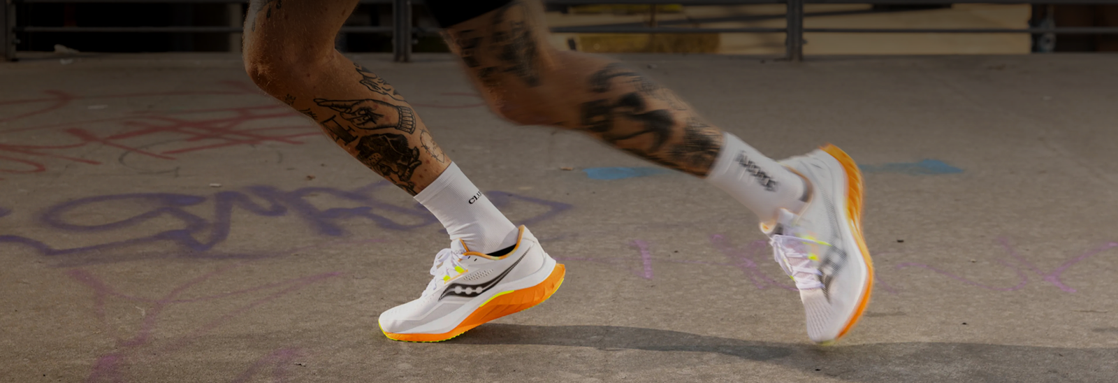 En person med tatoverede ben løber på et fortov, der er dækket af graffiti, iført hvide løbesko med orange såler og hvide sokker.