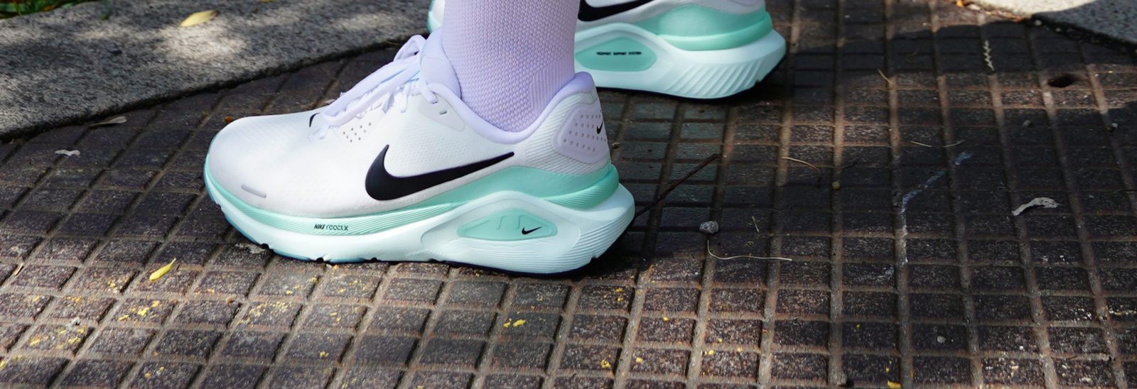 Nærbillede af en person iført hvide og mintgrønne Nike-løbesko med sorte swooshes, der står på et flisebelagt fortov udendørs. En parkscene med træer og mennesker er svagt synlig ovenover.