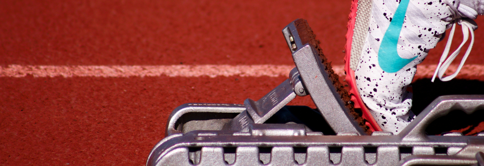 Nærbillede af en persons fod i en hvid løbesko med blå swoosh og røde prikker, placeret i en startblok på en rød bane.