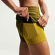 En kvinde iført Nike Nk Swift Df Mr 2N1 Shrt løbeshorts i Olive Flak/Peat Moss lægger en smartphone i sidelommen; shortsene har Dri-FIT-teknologi og er vist på en almindelig, lys baggrund.