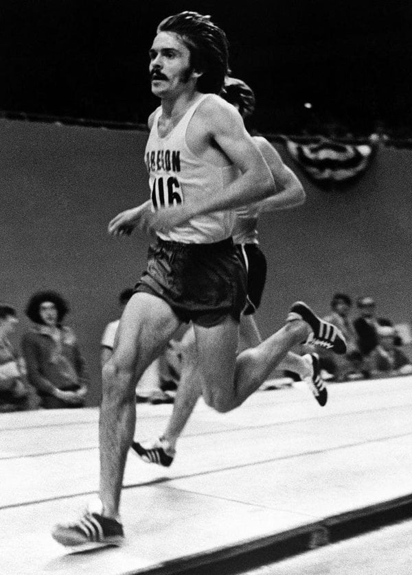 En mandlig løber med overskæg og langt hår, iført en Oregon-tanktop og shorts, løber indendørs på en bane med tilskuere siddende i baggrunden.