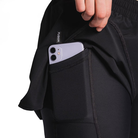 En person stikker en smartphone ned i sidelommen på et par sorte leggings med synlige sømme og kombinerer dem perfekt med Fusion Run Shorts, Dame, Black for maksimal komfort og bekvemmelighed.