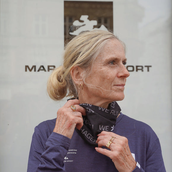 En kvinde med blondt hår i hestehale står foran et butiksvindue iført en blå langærmet bluse og »Marathon Sport Multiscarf, Unisex, Sort« fra Marathon Sport med »WE RUN«-designet. Hun kigger til siden, mens hun holder multiscarfet tæt ved halsen, hvilket fremhæver dette stilfulde løbetilbehør – ideelt til aktive dage.