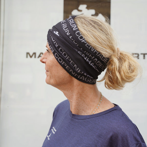 En person med blondt hår sat op i en lav, rodet knold bærer et sort »Copenhagen«-hårbånd og en blå skjorte og står foran en hvid baggrund, hvor en del af teksten er synlig. Deres look fremhæves af Marathon Sport Multiscarf, Unisex, Sort fra Marathon Sport – ideel til en aktiv livsstil eller som en del af det uundværlige løbetilbehør.