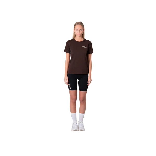 En person står på en hvid baggrund iført en Liiteguard Ground-Tech T-shirt, Dame, Coffee Bean, sammen med sorte shorts, hvide strømper og hvide sneakers. De ser fremad med armene ned langs siden.