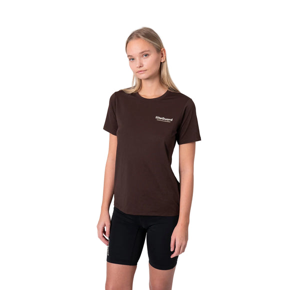 En kvinde med glat, skulderlangt blondt hår og et neutralt udtryk står på en hvid baggrund iført en Liiteguard Ground-Tech T-shirt, Dame, Coffee Bean med S.Café-materiale og et lille hvidt logo på brystet, sammen med sorte shorts.