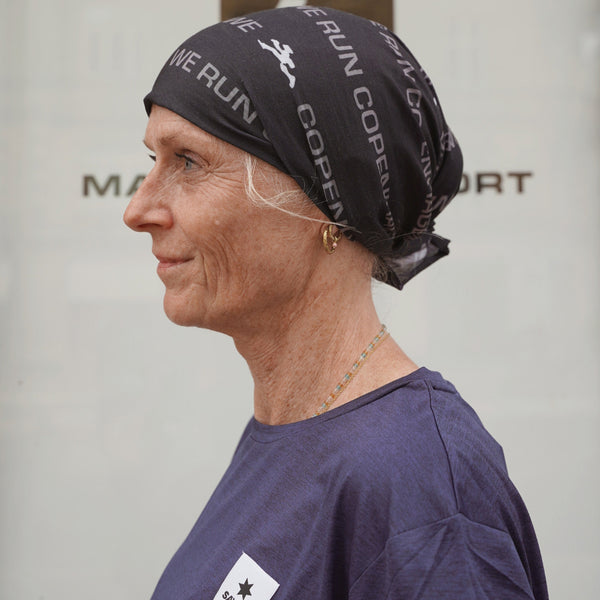 En lyshudet ældre kvinde set i profil bærer et sort »Marathon Sport Multiscarf« fra Marathon Sport og en lilla T-shirt. Hun har kort gråt hår og guldringe i ørerne. Baggrunden er neutral og let sløret.